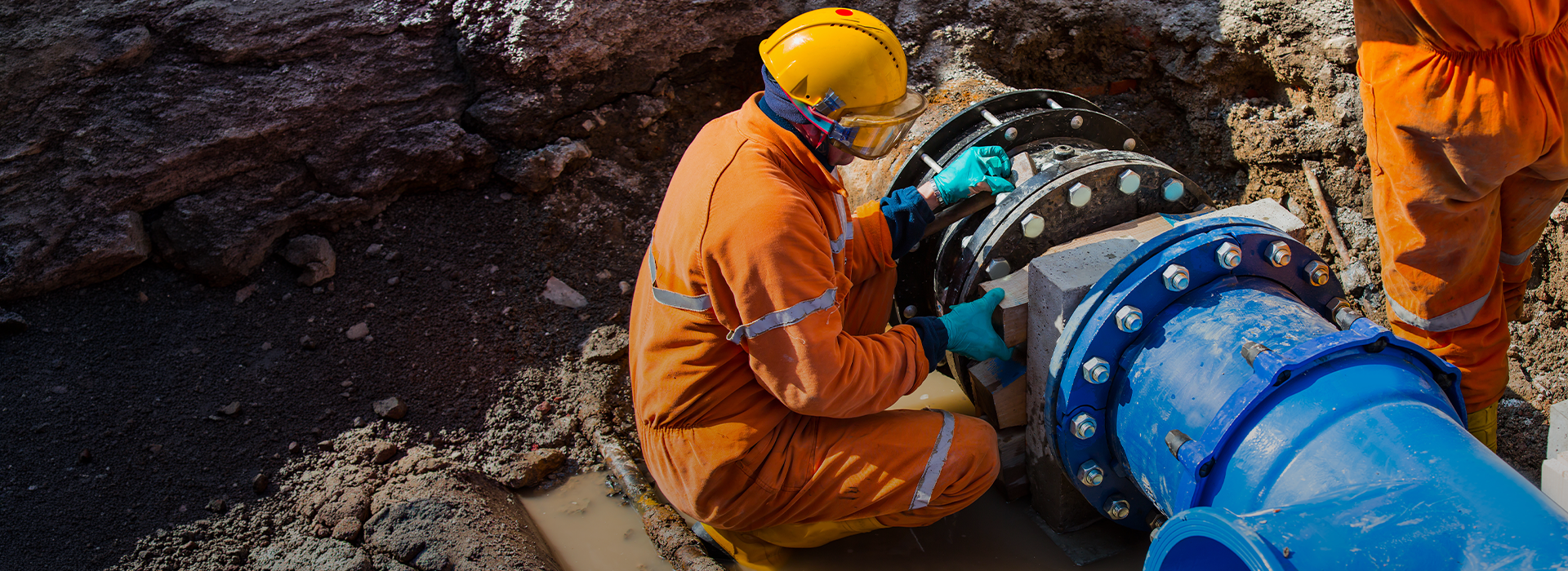 Zwei Bauarbeiter in orangenen Anzügen an einem großen Wasserrohr in einem Erdloch